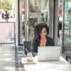 High angle of pensive African American female freelancer in glasses and casual clothes focusing on screen and interacting with netbook while sitting at table with glass of yummy drink on cafe terrace in sunny day